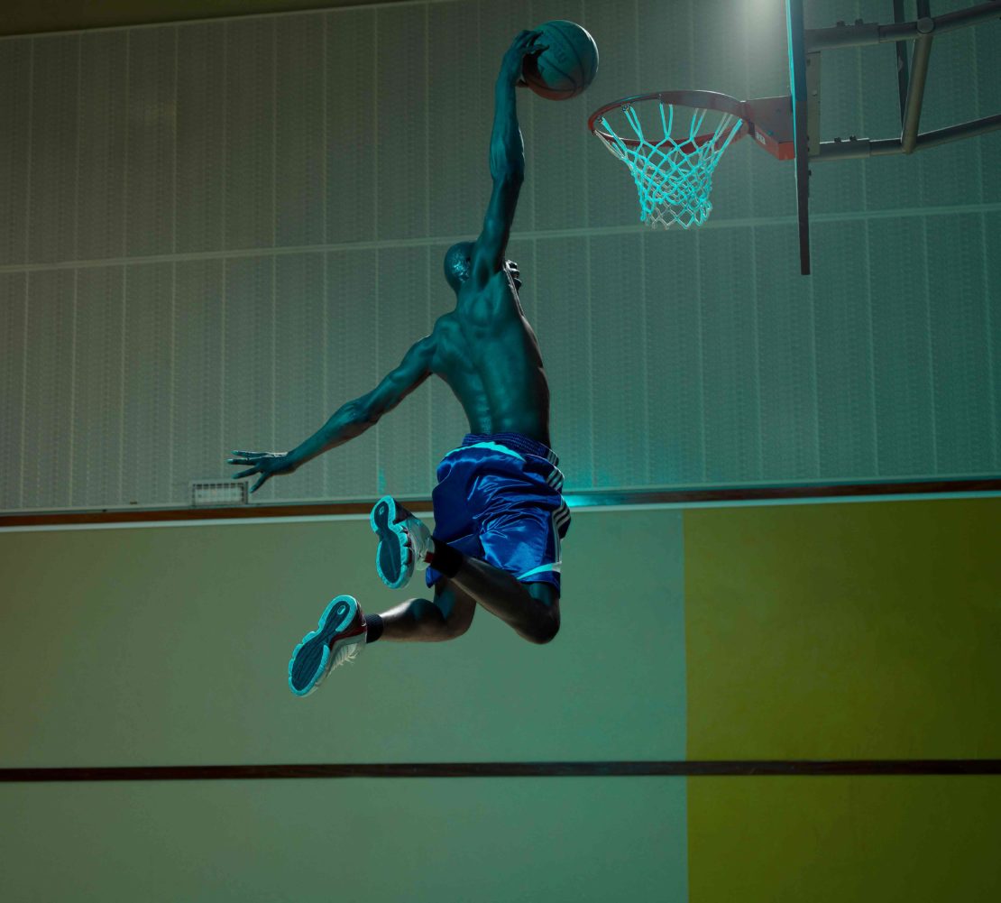 A shirtless basketball player in mid-air with a ball, poised to dunk, with dramatic turquoise lighting and a green-toned court.