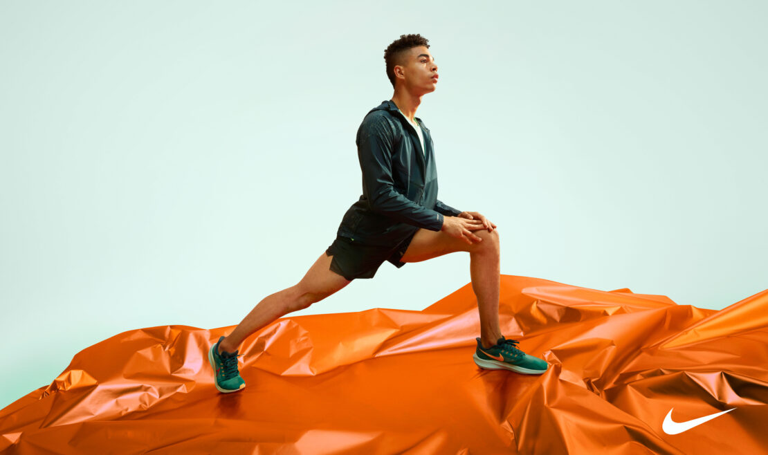 Athletic man in black athletic wear performs a leg stretch on an orange, wrinkled surface against a light blue background.