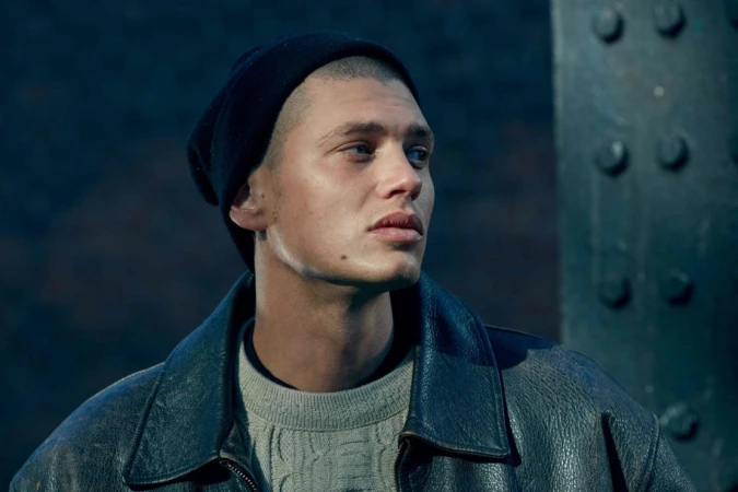 Close-up portrait of a young man wearing a black beanie, cream knit sweater, and dark leather jacket against a steel bridge structure.
