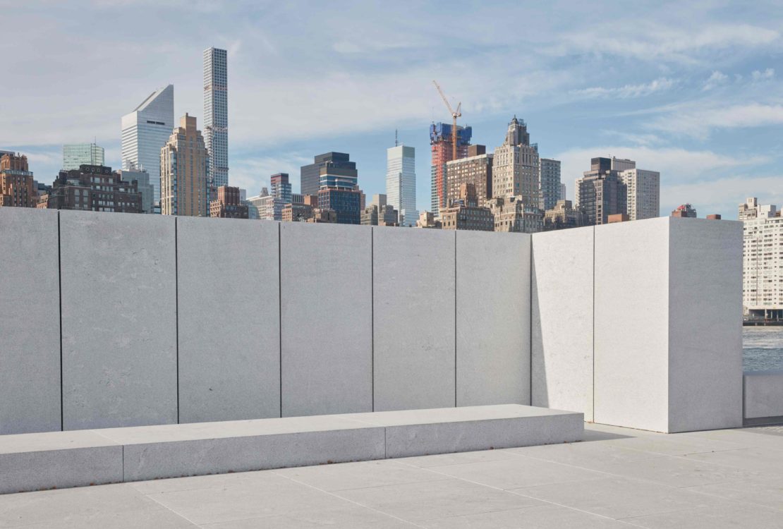 Concrete structure with a bench overlooks a cityscape with multiple skyscrapers under a partly cloudy blue sky during daylight hours.