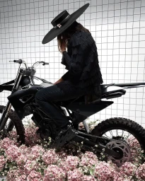 Model wearing a wide-brimmed black hat and ruffled black top seated on a dark motorcycle surrounded by pink flowers.