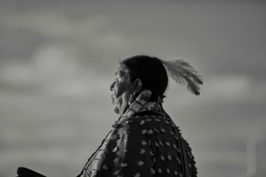 Profile portrait of an elderly individual wearing traditional regalia with a feather in dark hair against an overcast sky.