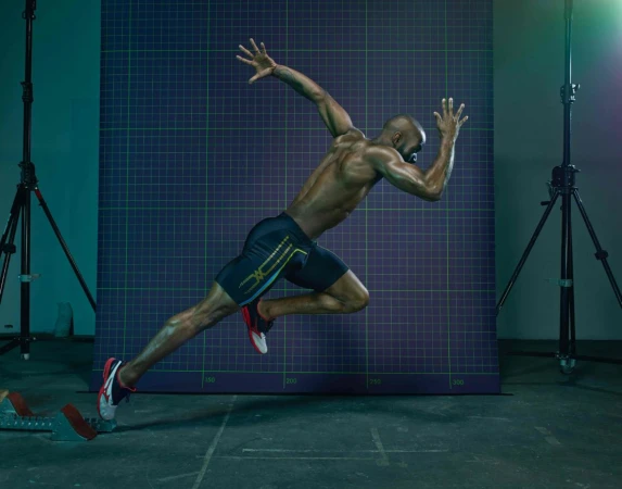 Black male athlete sprinting in mid-air against a purple grid backdrop with studio lighting and light stands