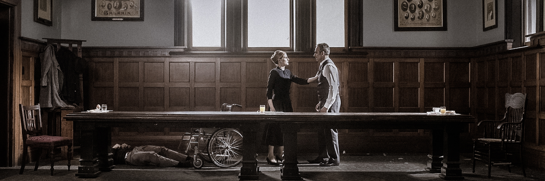 Indoor shot of a vintage-styled room with a man and woman standing next to a long wooden table, with a man lying underneath.