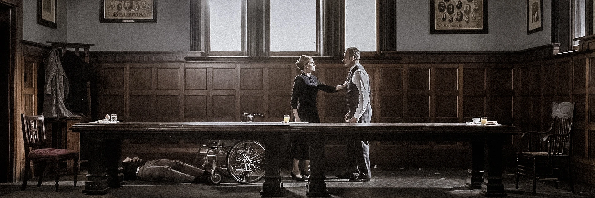 Indoor shot of a vintage-styled room with a man and woman standing next to a long wooden table, with a man lying underneath.