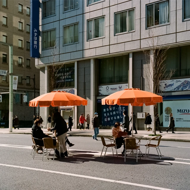 Outdoor cafe seating on an urban street with orange umbrellas, surrounded by pedestrians and modern buildings on a sunny day.