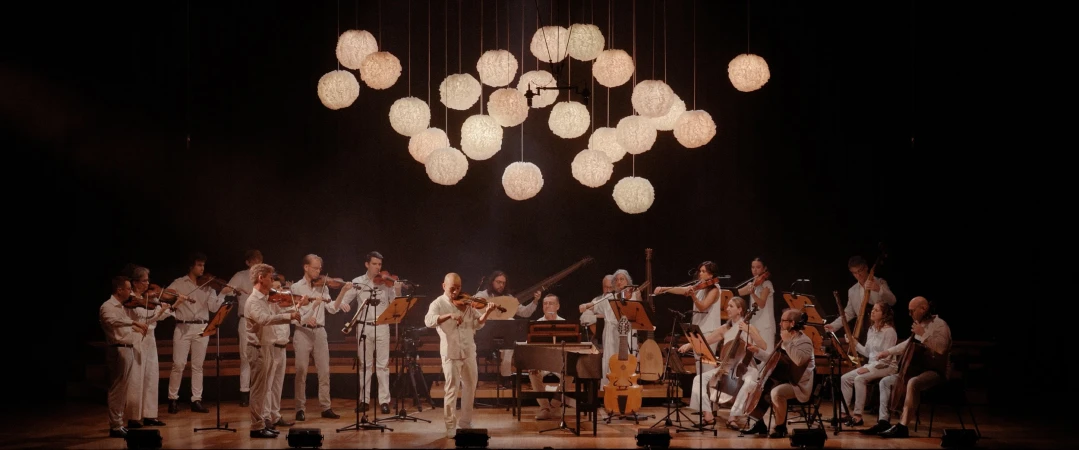 Orchestra performing on stage under glowing lights. Musicians play violins and other instruments in a dimly lit concert hall setting.