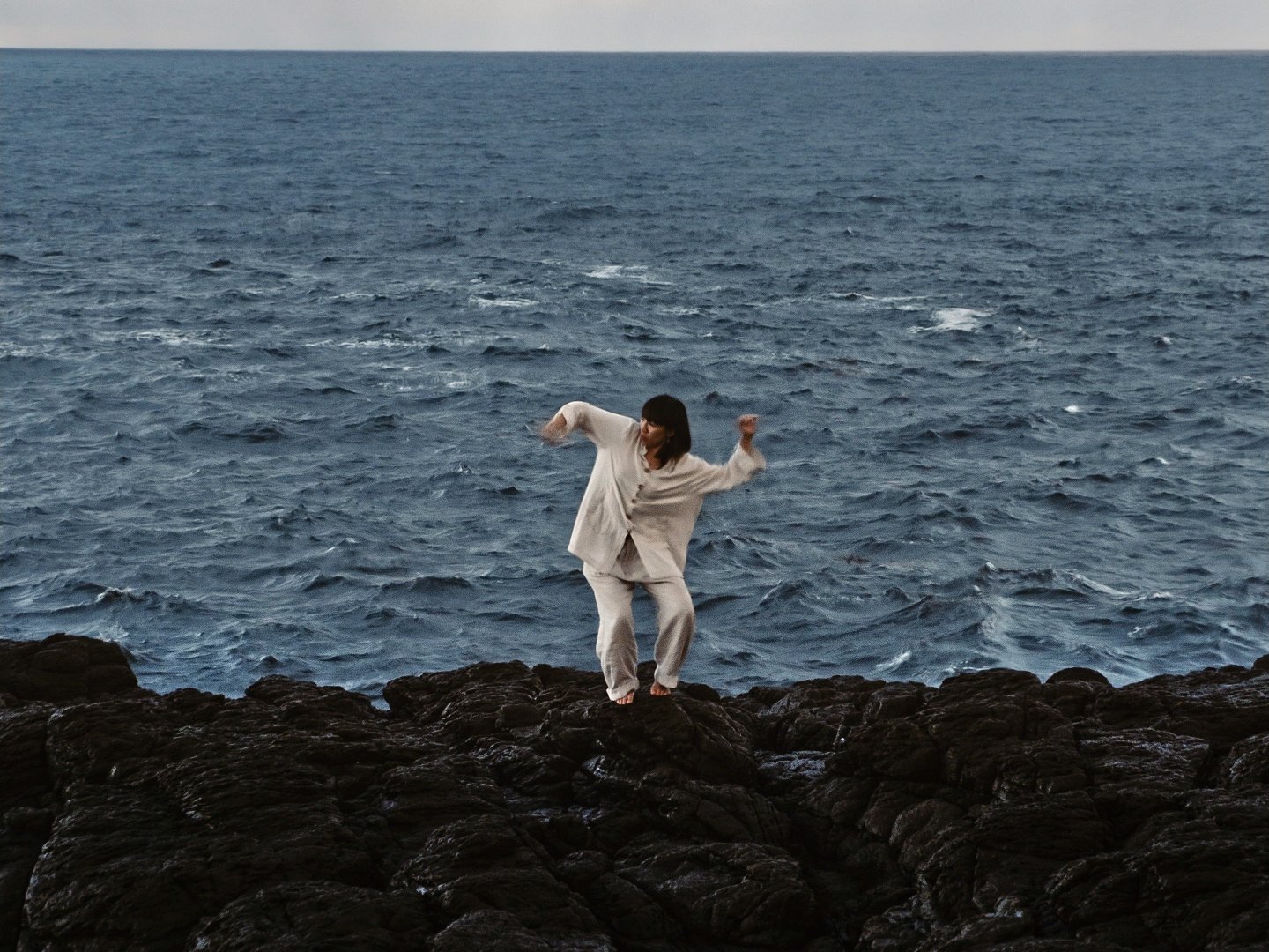 Adult stands on black rocky shore with waving blue sea in background and overcast sky, wearing off-white linen suit.
