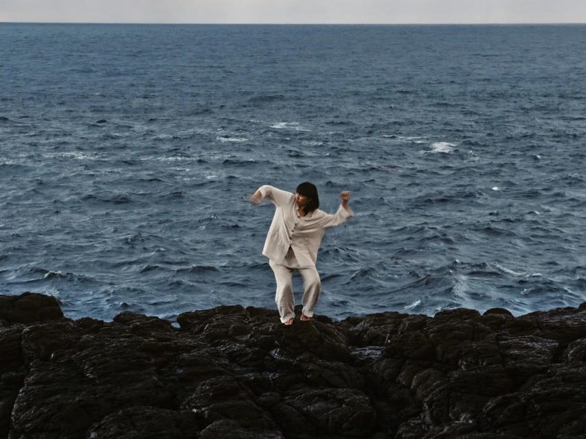 Adult stands on black rocky shore with waving blue sea in background and overcast sky, wearing off-white linen suit.