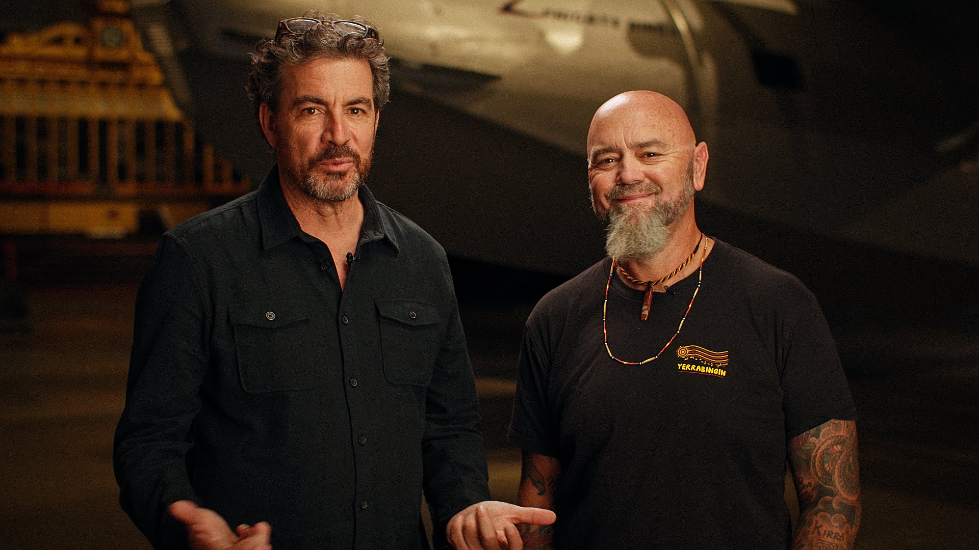 Two adult men stand in front of a dark background with industrial equipment; they are wearing casual clothing and looking at the camera.