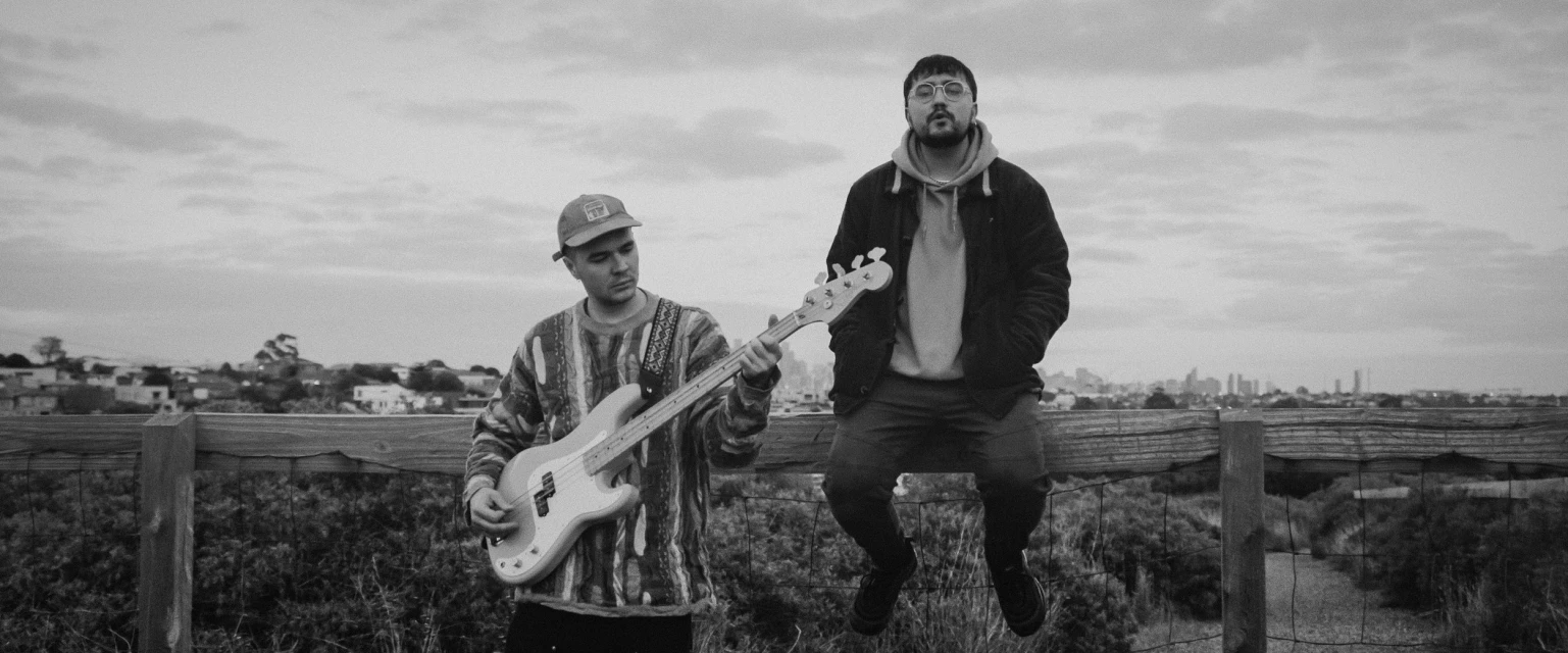 Black and white shot of two adult males near a wooden fence; one man plays a guitar, and the other sits with a city skyline in the background.