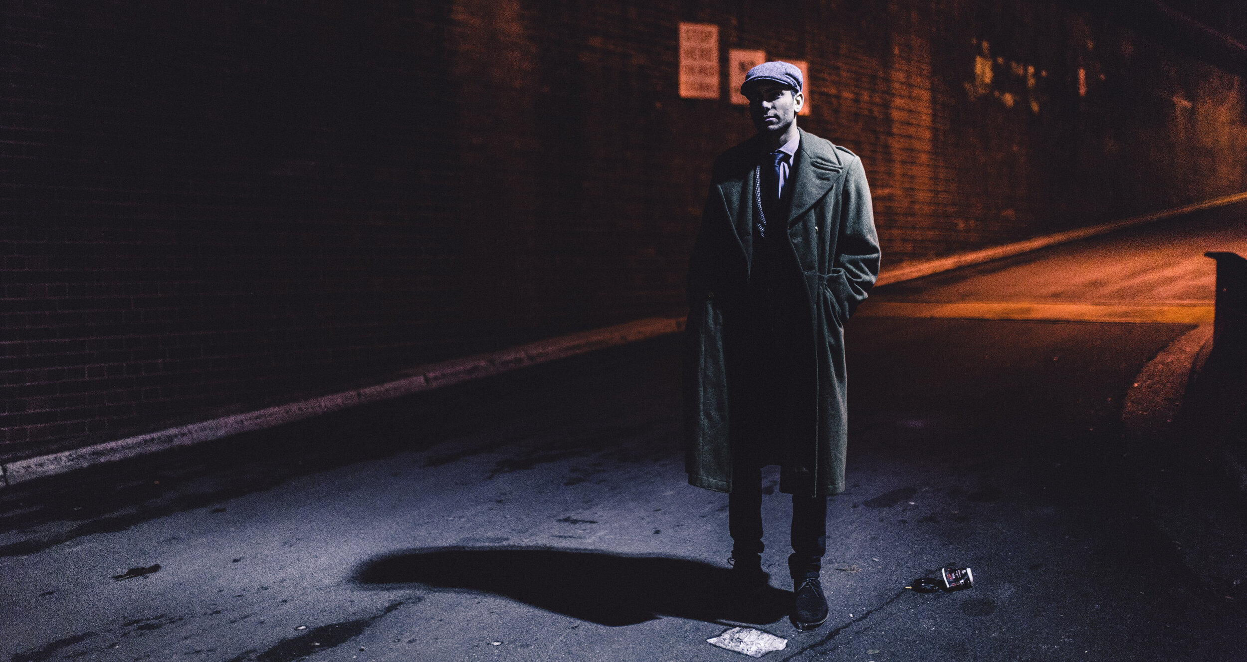 Man in an overcoat and newsboy cap stands on a dark street with brick walls under low light, casting shadows on the ground.