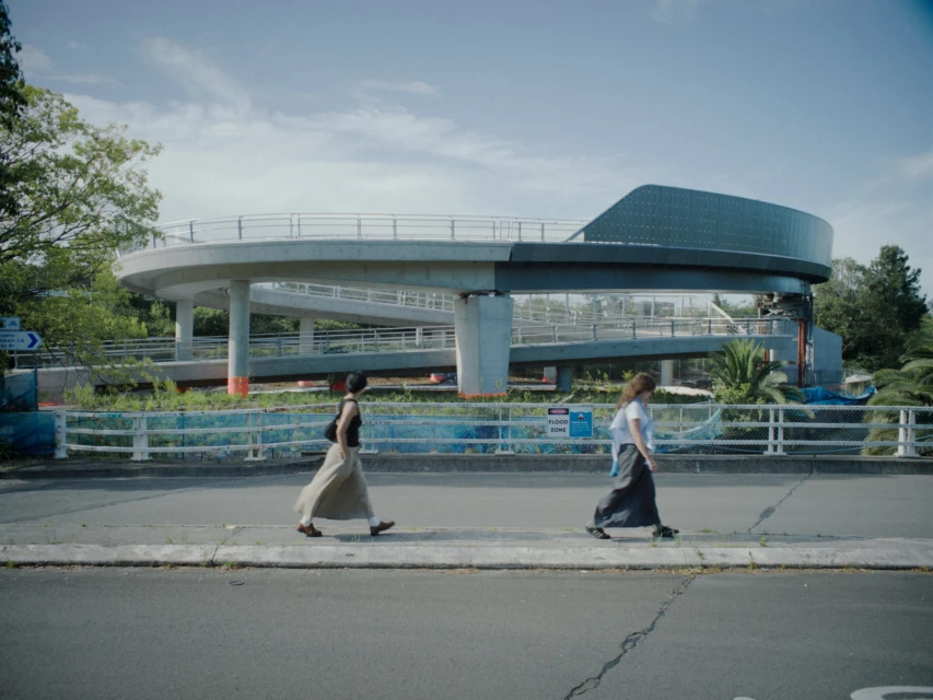 Two women walking side-by-side on a city street with an elevated walkway and lush trees under a bright, sunny sky.