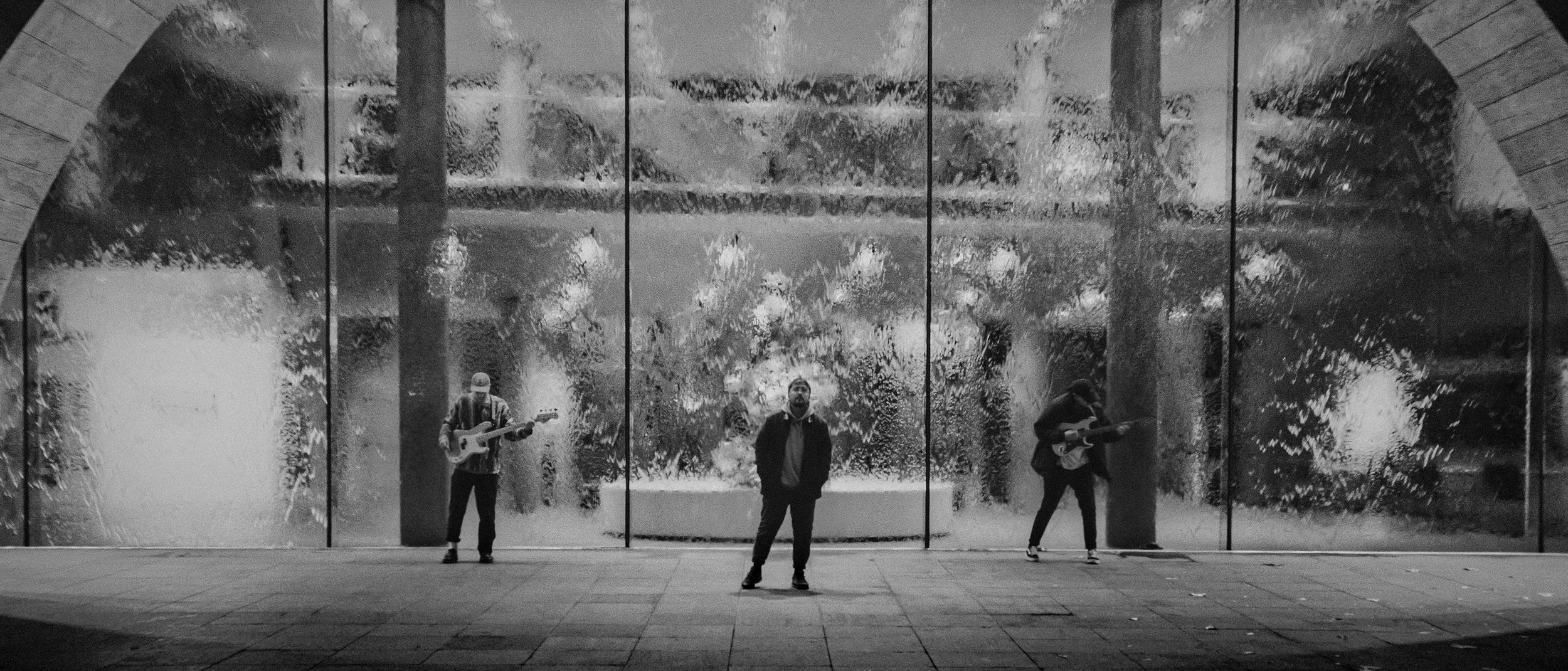 Three musicians stand in front of a waterfall wall, two playing electric guitars in a monochromatic urban setting.