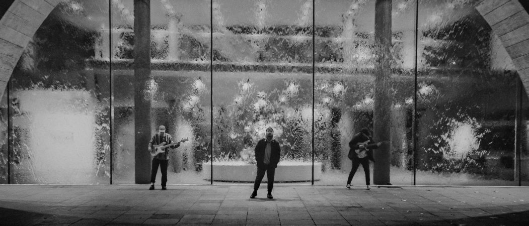 Three musicians stand in front of a waterfall wall, two playing electric guitars in a monochromatic urban setting.
