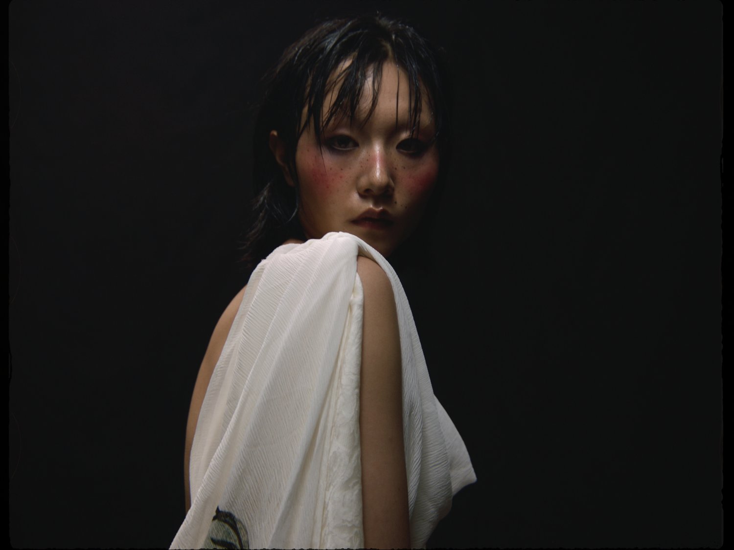 Fashion portrait of an Asian adult woman with heavy makeup and draped in a white textured fabric, set against a dark black background.