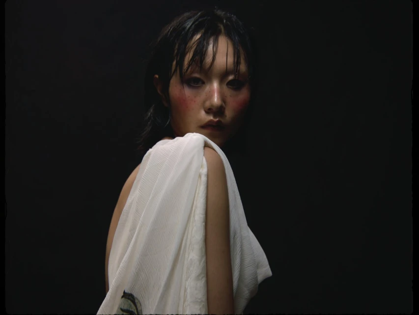 Fashion portrait of an Asian adult woman with heavy makeup and draped in a white textured fabric, set against a dark black background.