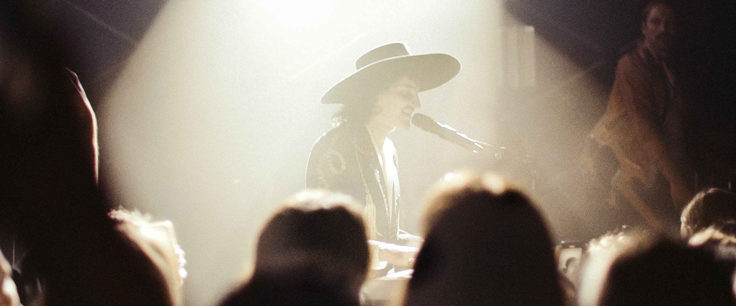 Singer in hat performs on stage under bright lights in front of audience members at a concert venue during an evening performance.