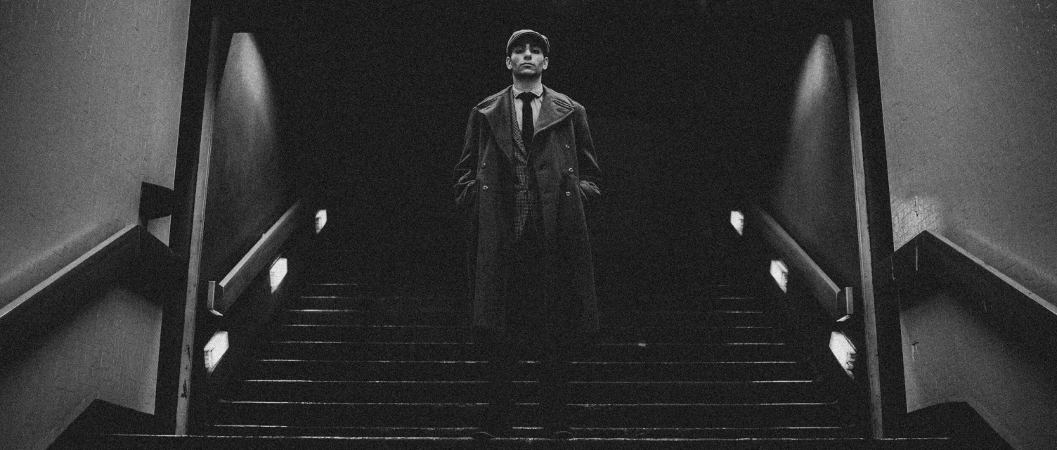 Monochrome portrait of a man in a coat and cap standing on stairs with illuminated handrails, looking toward the viewer.