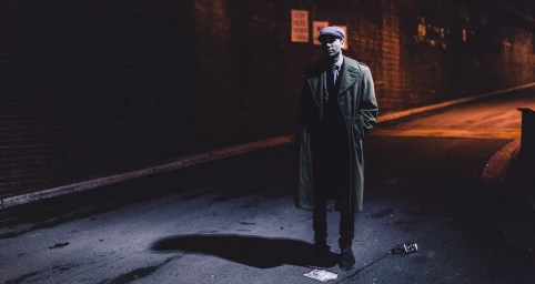 A man in a long green coat and flat cap stands on an asphalt alley under dim overhead lighting with a long shadow cast behind him.