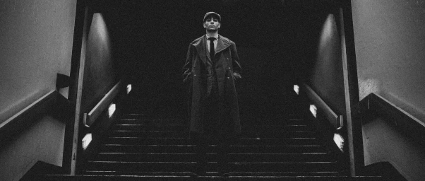 Man wearing a long coat and flat cap standing on stairs in a gritty monochrome setting by Tom Pennell