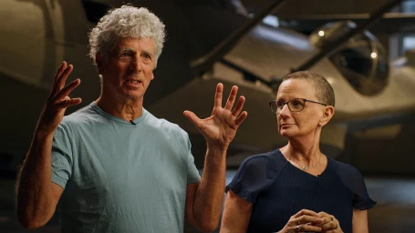 Older man with curly white hair wearing a light blue t-shirt gesturing while standing beside a woman with glasses