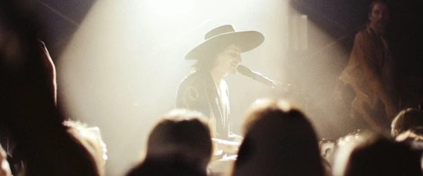 Performer wearing a wide-brim hat singing into a microphone on stage under a spotlight, framed by audience silhouettes.