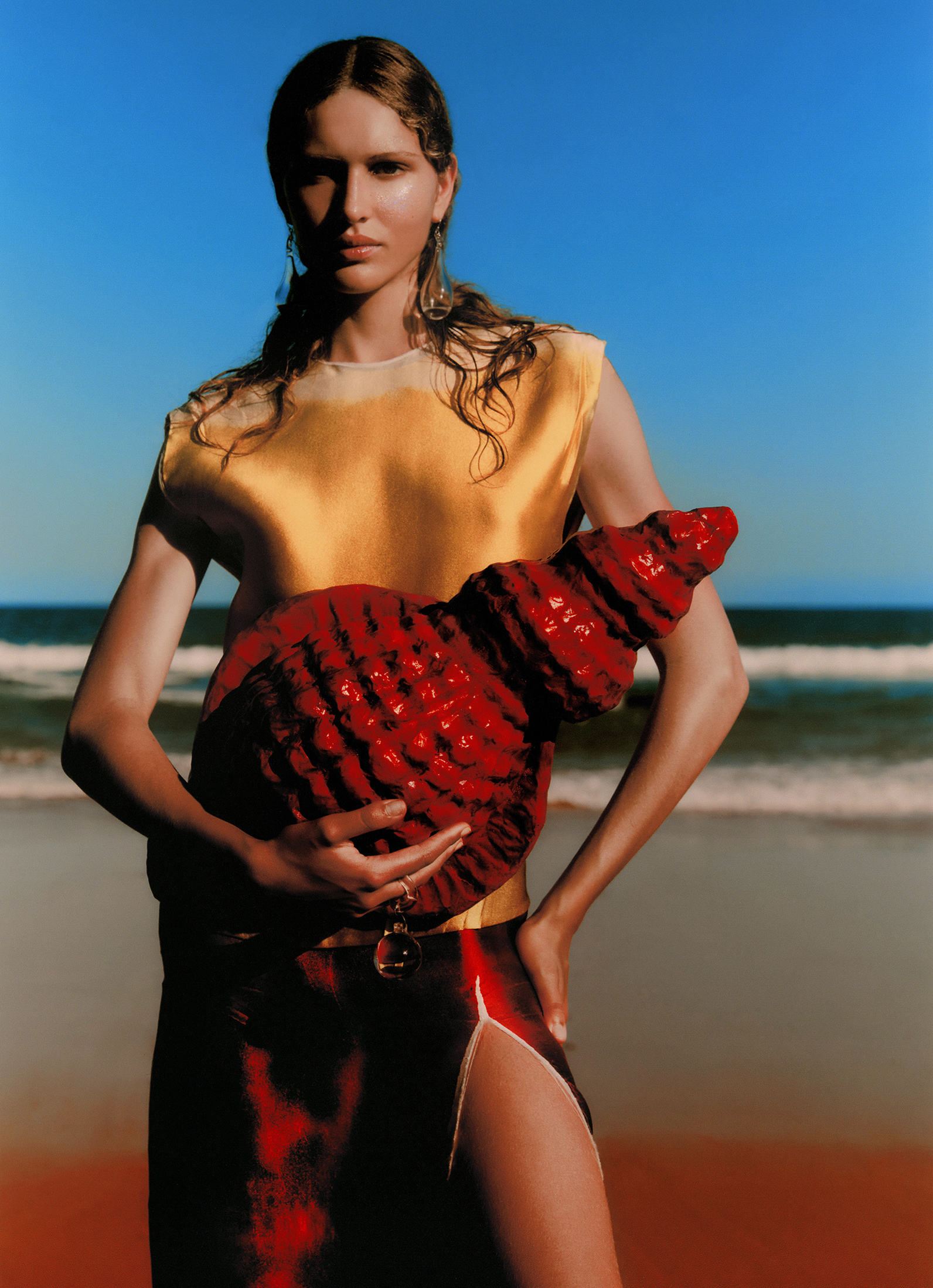Fashion shot of a woman posing on a beach, holding a red decorative object, wearing a yellow top and skirt, with blue sky backdrop.