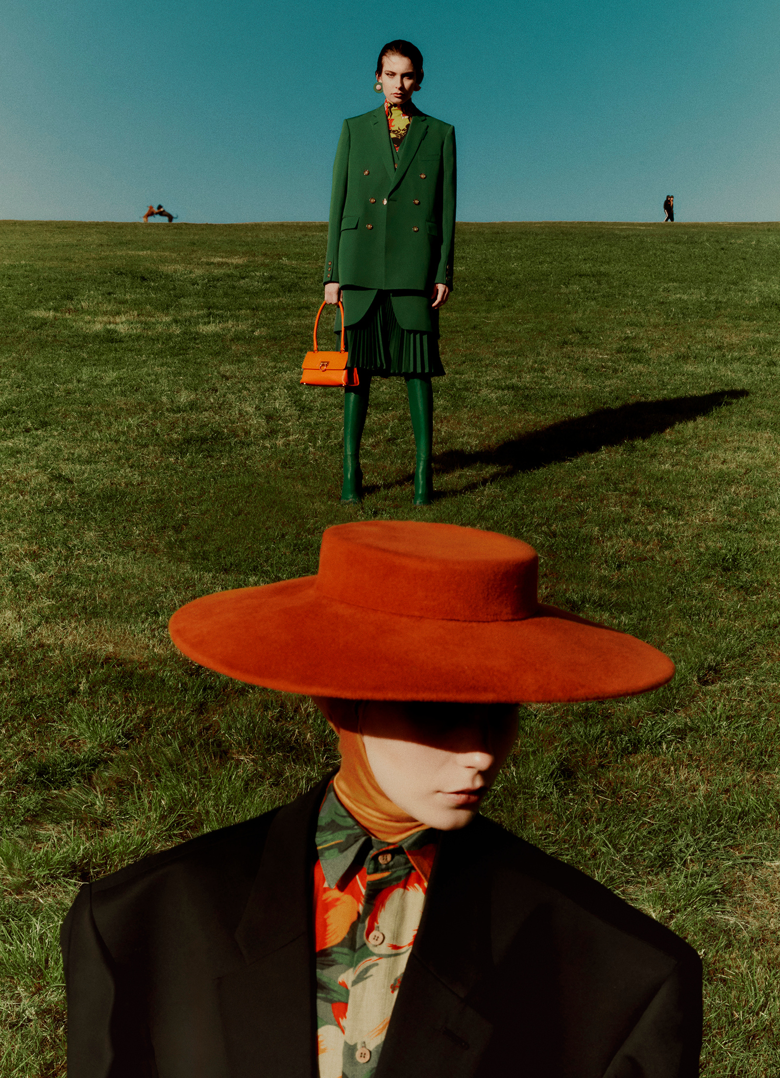 Two fashion models pose in a grassy field, one in a green suit with an orange bag, the other with an orange hat and floral shirt.