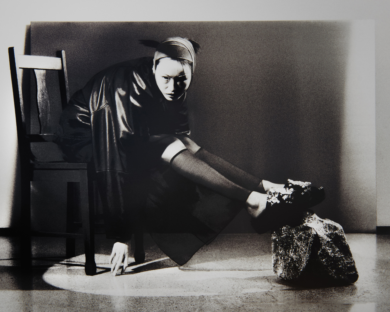 Black and white fashion photograph of a person in a headscarf and black leather outfit sitting on a chair indoors with a spotlight.