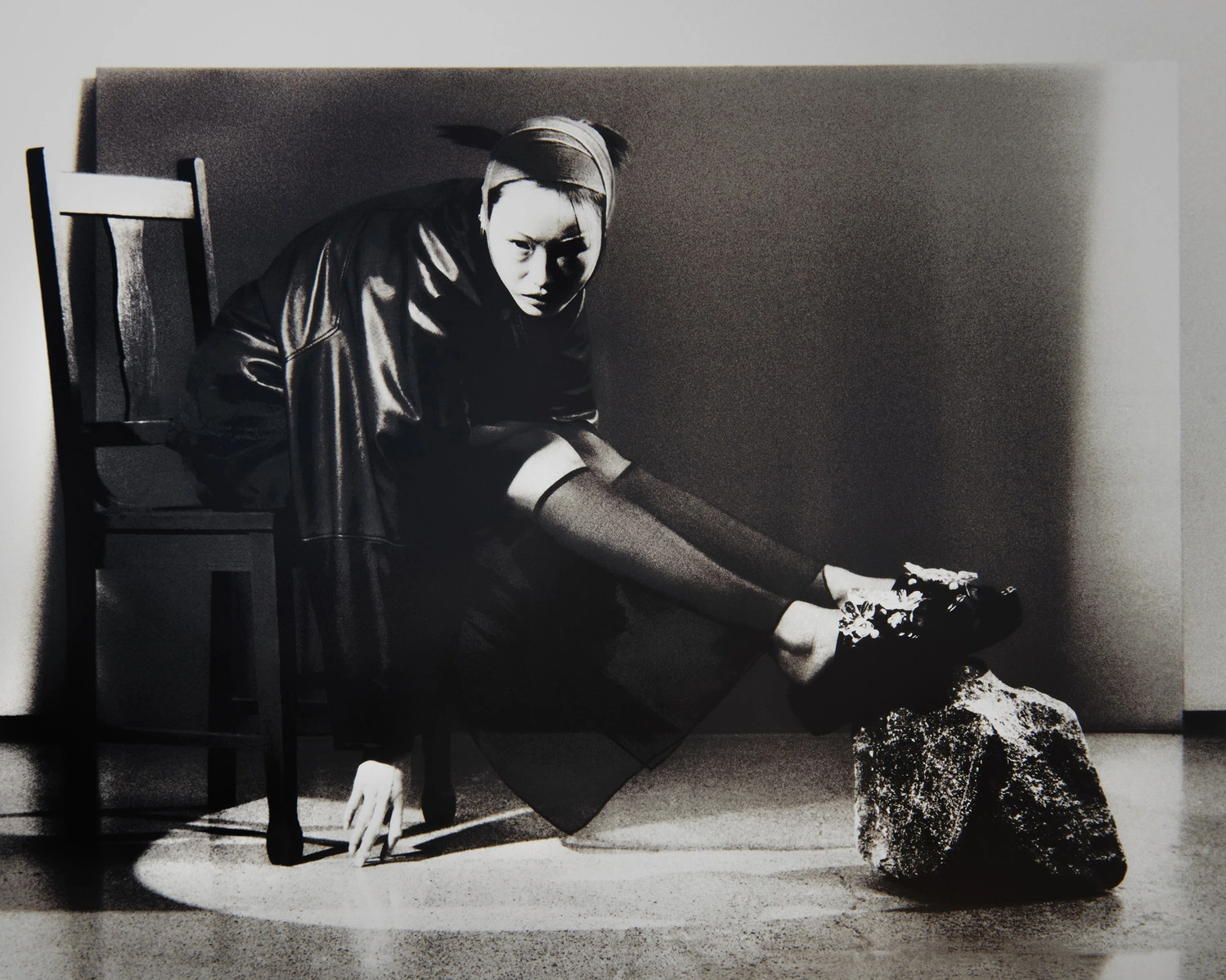 Model seated on a wooden chair wearing dark clothing and a headband with feet resting on a textured object in studio lighting