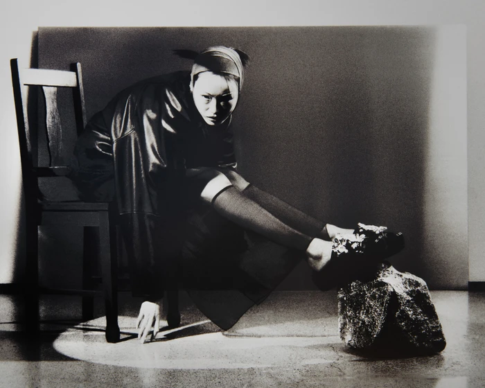 Model seated on a wooden chair wearing dark clothing and a headband with feet resting on a textured object in studio lighting