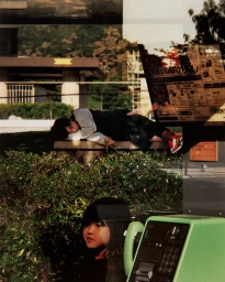 Reflective glass shot showing a child sleeping on a bench outdoors and a young person near a public payphone
