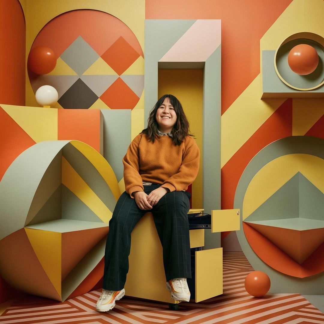 Smiling woman with dark hair wearing orange sweater seated on geometric yellow shelving, surrounded by geometric paper art props.