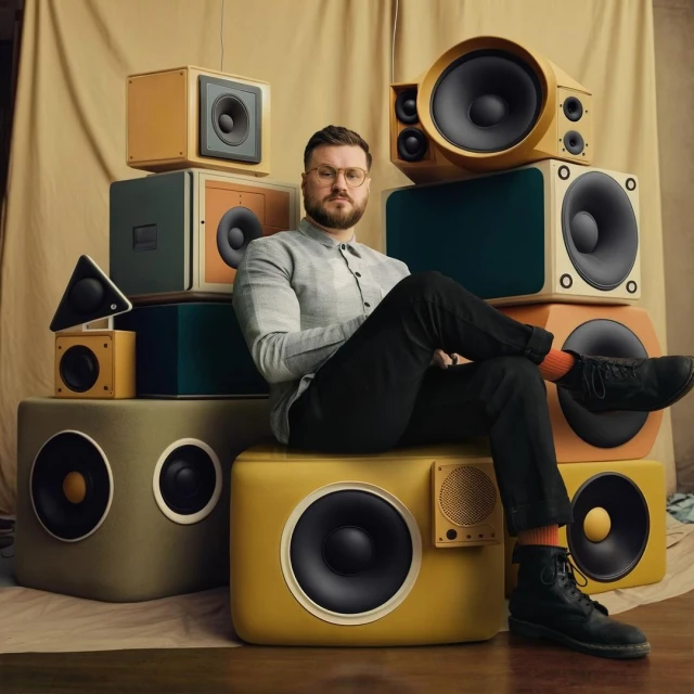 A man with glasses sits cross-legged on a stack of speakers in a photo studio with a beige background. He wears a dress shirt and black pants.