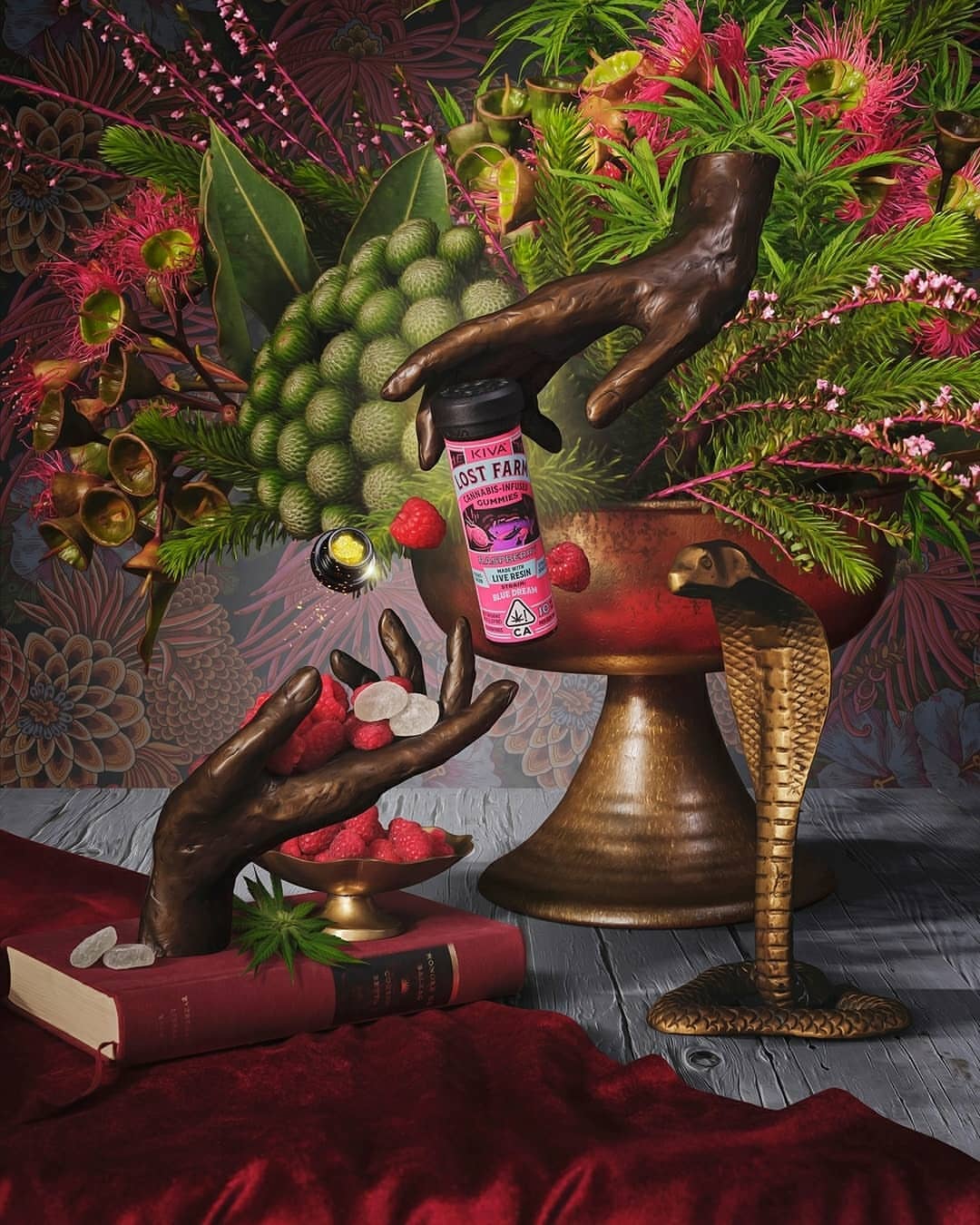 Still life featuring bronze hand sculptures, a jar of cannabis gummies, fruit, flowers, and books, creating a vibrant botanical scene.