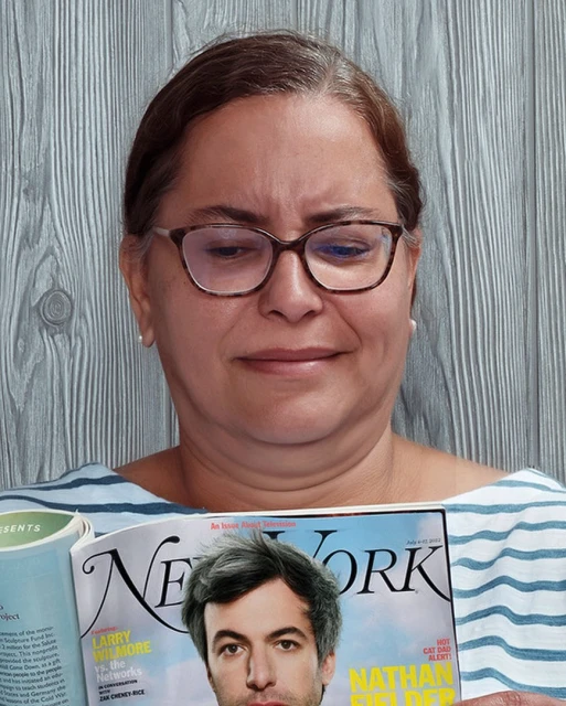 Adult female indoors holds a New York magazine with a man on the cover, wearing eyeglasses, and a striped shirt against a wood background.