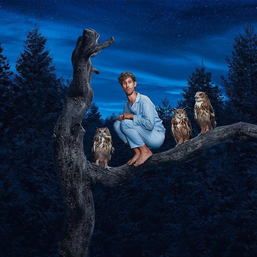Man squats barefoot on tree branch surrounded by owls under starry sky, wearing pajamas, in a nighttime forest scene.