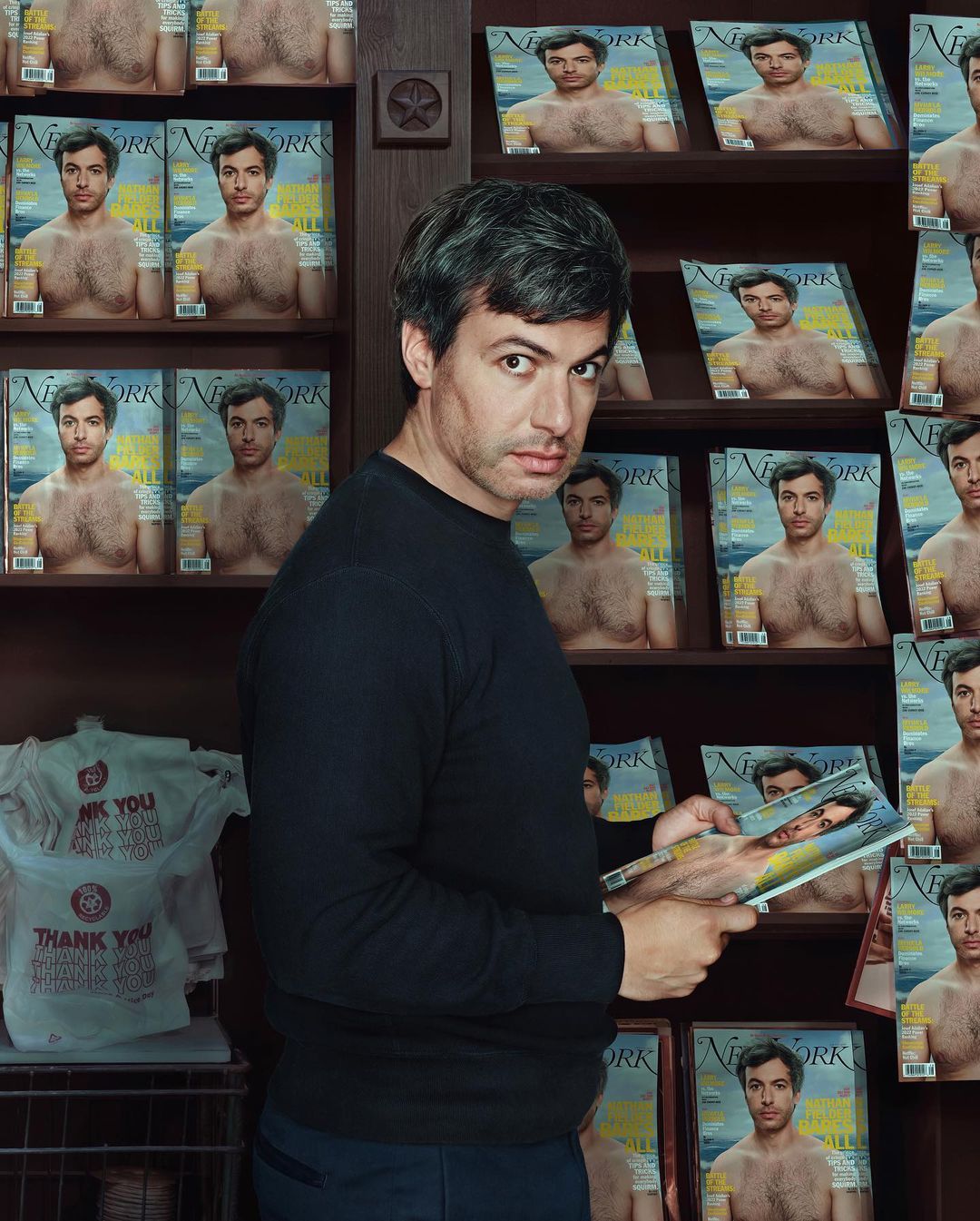 Man in a black long-sleeve shirt stands beside a bookshelf filled with magazines, one of which he holds, in an indoor setting.