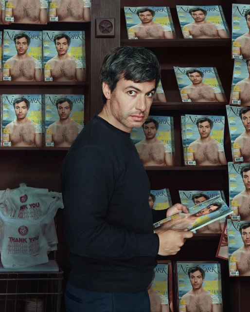Man in a black long-sleeve shirt stands beside a bookshelf filled with magazines, one of which he holds, in an indoor setting.