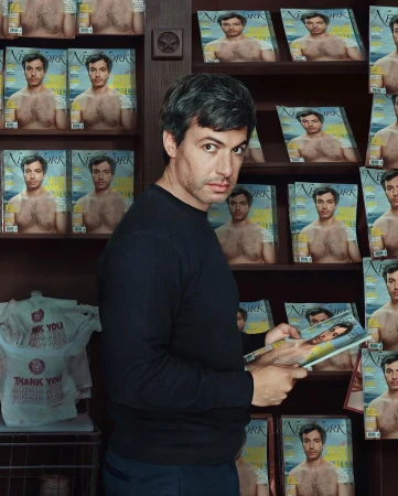 Man in a black long-sleeve shirt stands beside a bookshelf filled with magazines, one of which he holds, in an indoor setting.