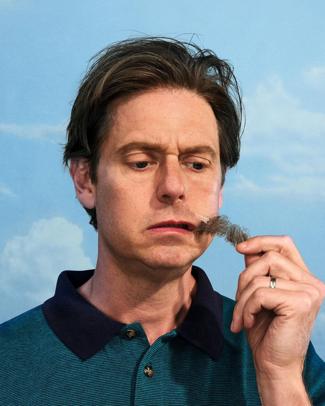 Portrait of a middle-aged man in a teal polo shirt holding a clump of dark facial hair towards his face, set against a blue sky backdrop.