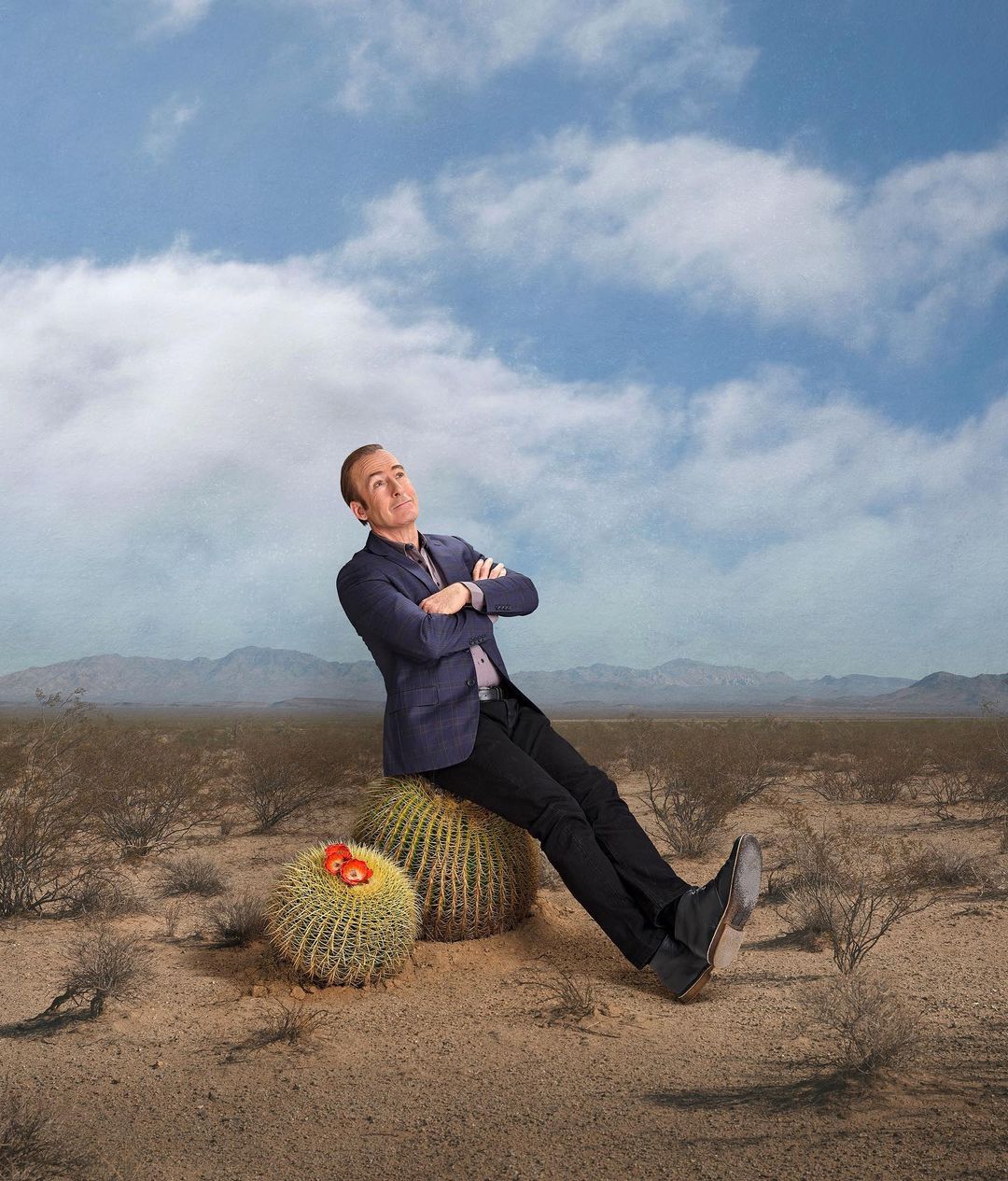 Adult male poses leaning on cactus in desert setting under blue, cloudy sky; wearing a navy jacket, black pants, and black boots.