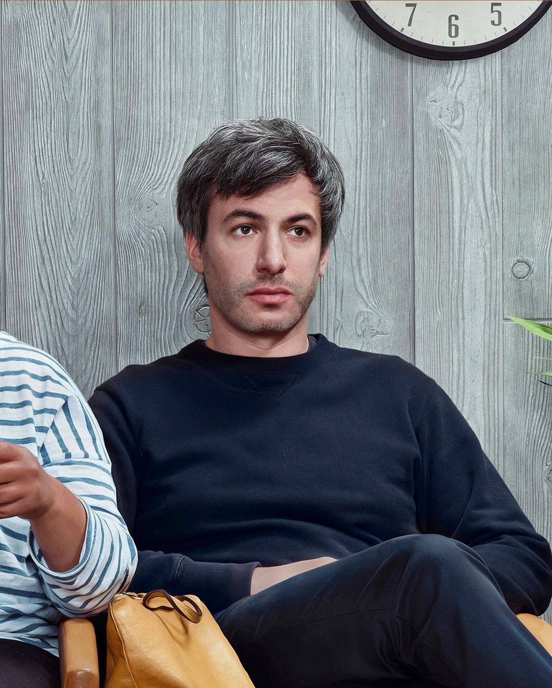 Adult male with grey hair sits on a tan chair indoors wearing a navy sweatshirt. Background includes a clock and grey wood paneling.