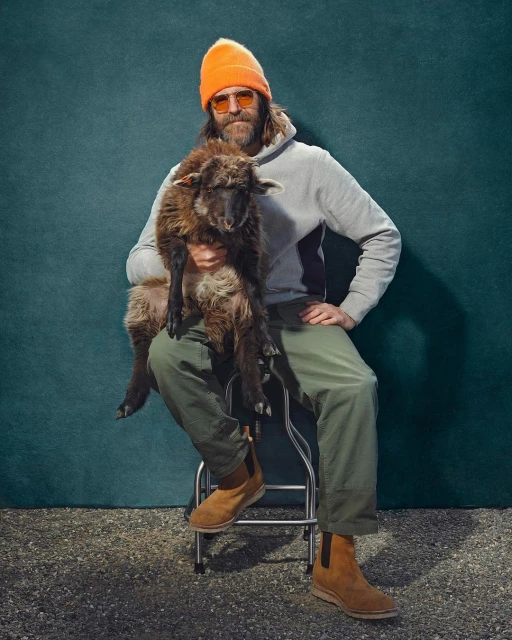 Man in grey sweatshirt and orange hat sits holding lamb against teal backdrop, wearing tan boots and orange sunglasses.