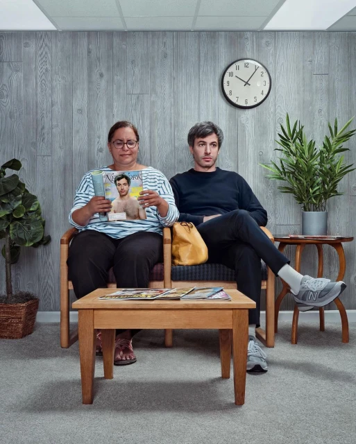 Two people sit in a waiting room with wood paneling, reading magazines and looking forward. Indoor setting, soft light, neutral colors.