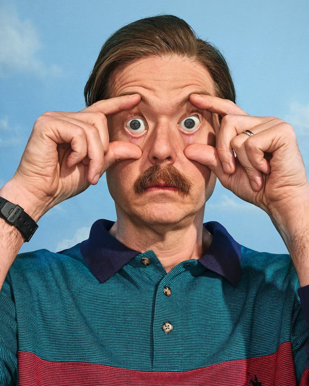 Man with mustache stretches his eyes open wide with his fingers; he wears a striped polo shirt against a bright blue sky.