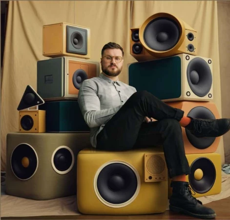 Man with beard and glasses sits on stack of retro speakers in studio with tan backdrop, wearing grey shirt, dark pants, black boots.