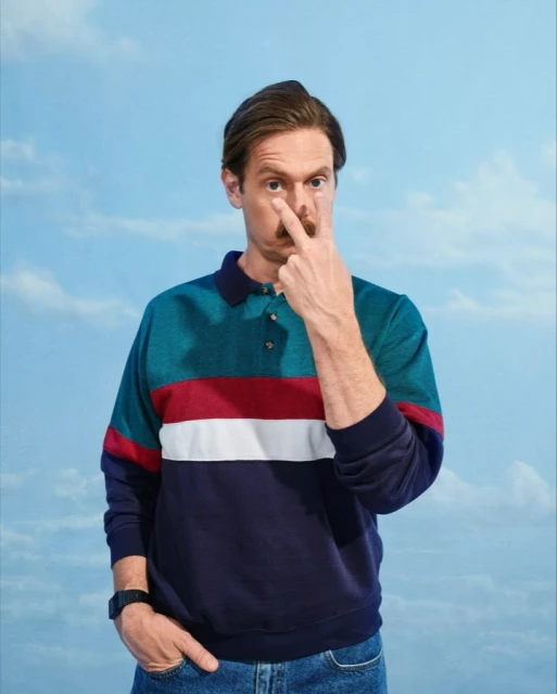 Portrait of a man in a blue and green color-blocked polo shirt pointing to his eye, set against a light blue sky backdrop.