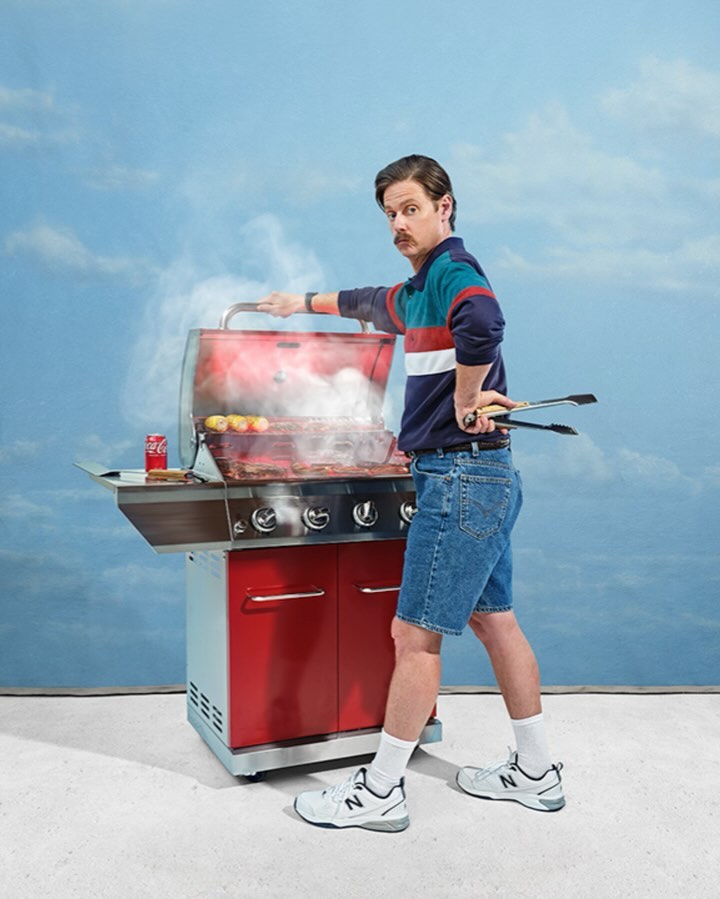 Man in a retro outfit grilling food on a red BBQ in front of a cloud-filled sky, wearing jean shorts, and holding metal tongs.